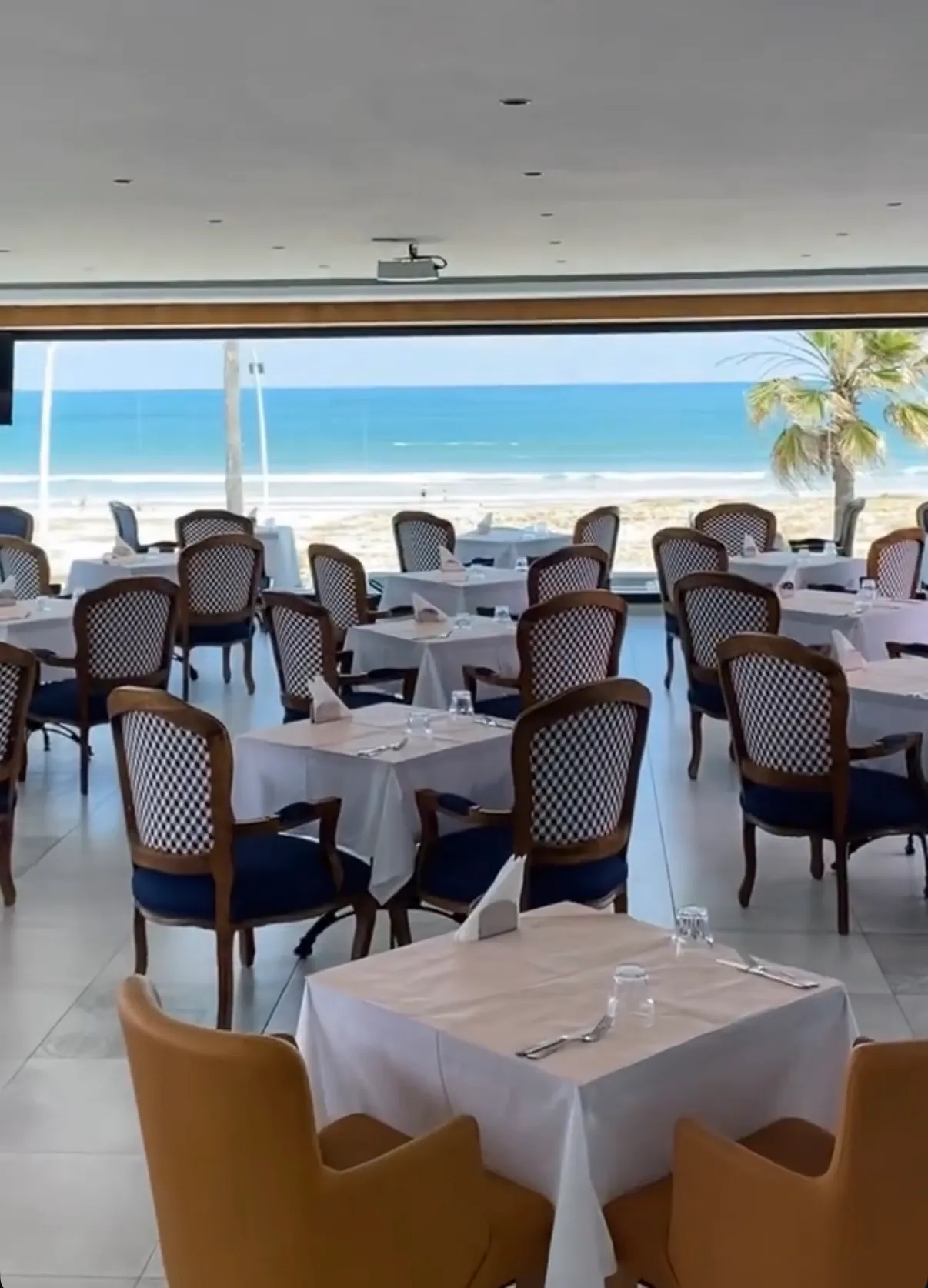 La salle de Mucho : tables nappées de blanc et fauteuils au dossier damier, face à la grande baie vitrée ouverte sur la plage et l'océan d'El Jadida