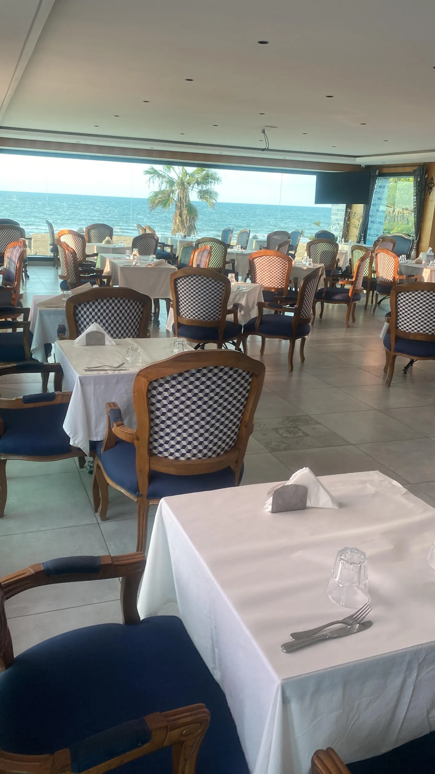 La salle de Mucho en journée : tables dressées, fauteuils au dossier damier et baie vitrée sur la plage et les palmiers
